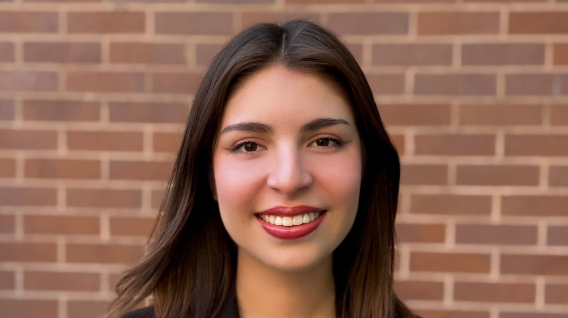Portrait of a current Gannon Scholar with long dark brown hair, smiling, standing in front of a brick wall.